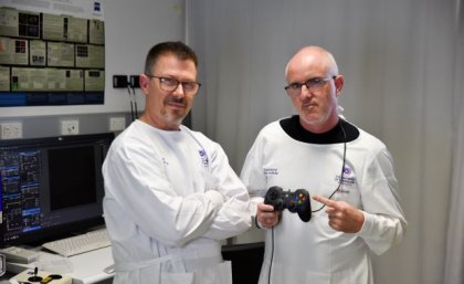 Two men in white lab coats in an office look at the camera, one holds a gaming controller. 